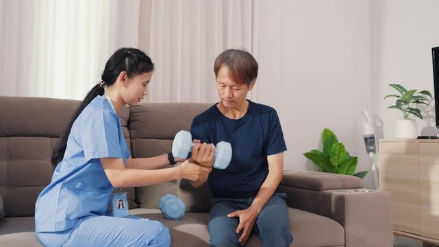 Supportive Asian therapist assisting male patient with dumbbell lifting exercise for physical therapy. Dedicated healthcare professional guiding strength recovery training on sofa.