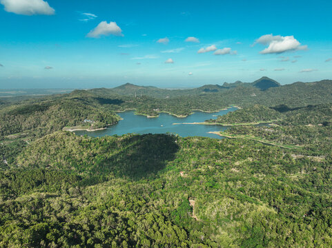 Aerial view of Sermo Reservoir Dam surrounded by lush green hills and dense tropical forest under a clear blue sky in Kecamatan Kokap, Daerah Istimewa Yogya, Indonesia.