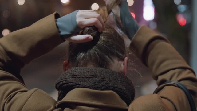 Night bus stop, white woman adjusts hair with gloved hands urban backdrop of blurred lights as she twists ponytail into neat bun, wearing warm coat and chunky scarf, purposeful rhythm suggests
