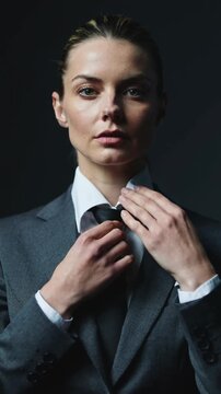 Woman in grey suit adjusting black necktie against dark studio background, medium shot