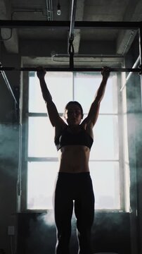 Athletic woman doing pull-ups in dark gym with bright window background and smoke, medium shot
