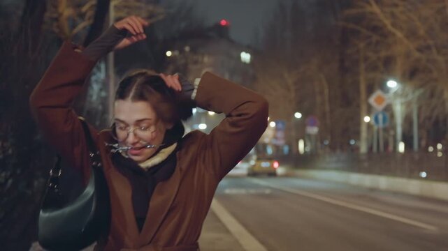 Night city sidewalk woman adjusting hair, bundled in brown coat and scarf, walking past glowing streetlights and taxis, candid urban portrait of a commuter tidying hairstyle with hands raised