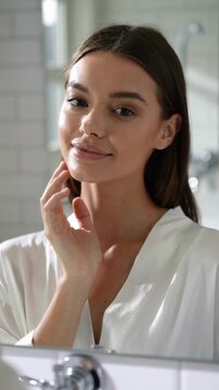 Young woman in white robe touching face in bathroom mirror, close up shot