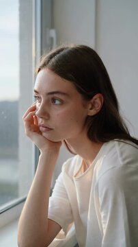 Young woman in white t-shirt resting chin on hand looking out window, medium shot, natural light