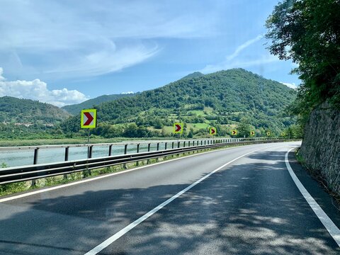 Carretera asfaltada con curva pronunciada bordeando un r&iacute;o y monta&ntilde;as verdes en un d&iacute;a soleado
