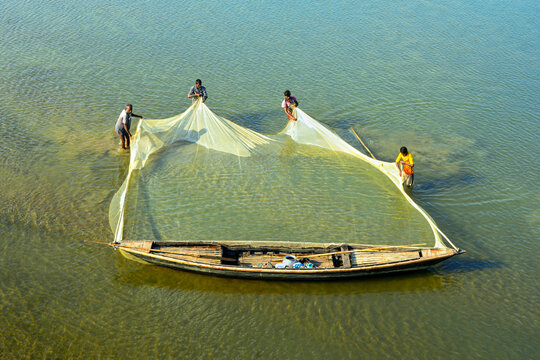 Aerial view of fishermen working together to pull a large fishing net from a wooden boat in the shallow water of Kurigram, Rangpur Division, Bangladesh.