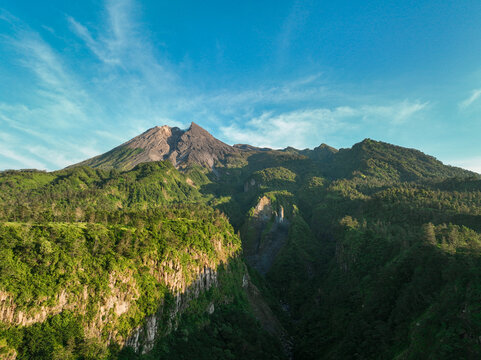 Aerial view of Merapi Mountain with rugged volcanic peak rising above lush green slopes and deep valleys under a clear blue sky in Kecamatan Cangkringan, Daerah Istimewa Yogya, Indonesia.