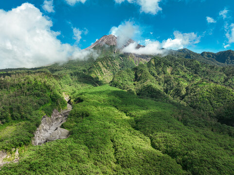 Aerial view of the lush green slopes of Mount Merapi with volcanic terrain, dense forest, and clouds under a bright blue sky in Kecamatan Cangkringan, Daerah Istimewa Yogya, Indonesia.