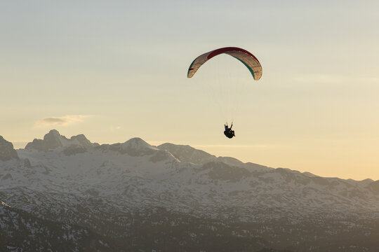 Aerial view of a paraglider soaring above the snow-capped Dachstein mountains during golden hour in Grobming, Styria, Austria.