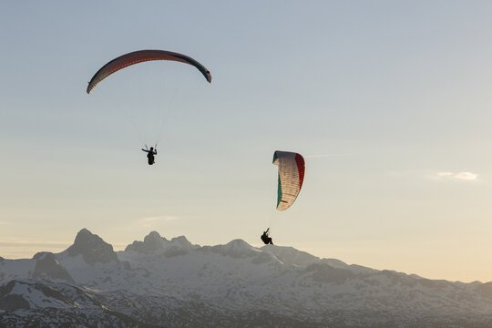 Aerial view of two paragliders soaring above the snow-capped Dachstein mountains during golden hour in Grobming, Styria, Austria.