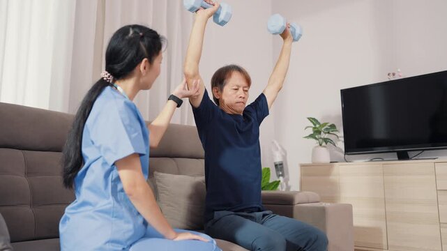Supportive Asian therapist assisting male patient with dumbbell lifting exercise for physical therapy. Dedicated healthcare professional guiding strength recovery training on sofa.