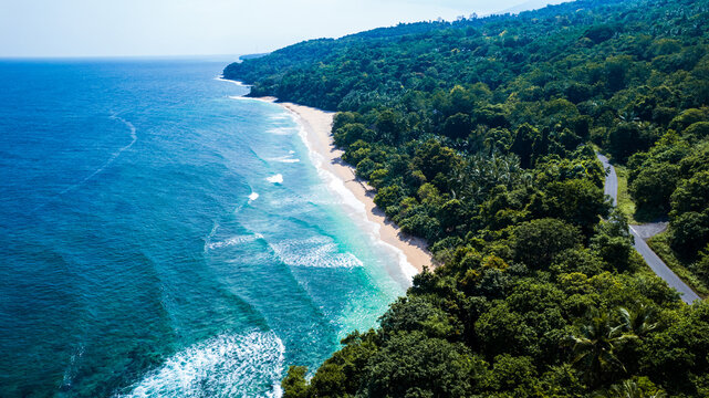 Aerial view of Waebela beach with turquoise waves crashing onto the sandy shore, bordered by dense tropical forest and a coastal road in Inerie, East Nusa Tenggara, Indonesia.
