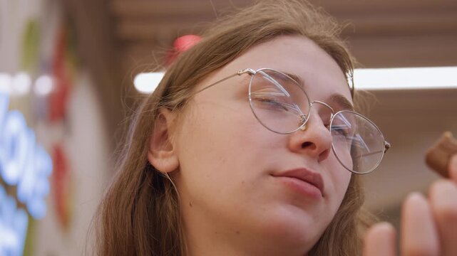 White woman tasting cookie in supermarket closeup shot wearing round glasses student shopper sampling pastry contemplative expression warm indoor lighting modern ceiling candid consumer moment