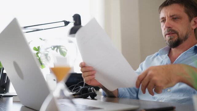 Focused businessman reviewing a business document while working at a laptop in his home office. Document analysis, business planning, professional decision making and remote work concept.