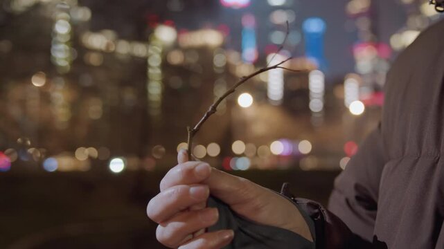 Closeup hands holding bud twig against city bokeh lights, finger touch and glove detail visible urban forager inspects tiny spring bud, moisture on branch and neon skyline blur suggest quiet hopeful