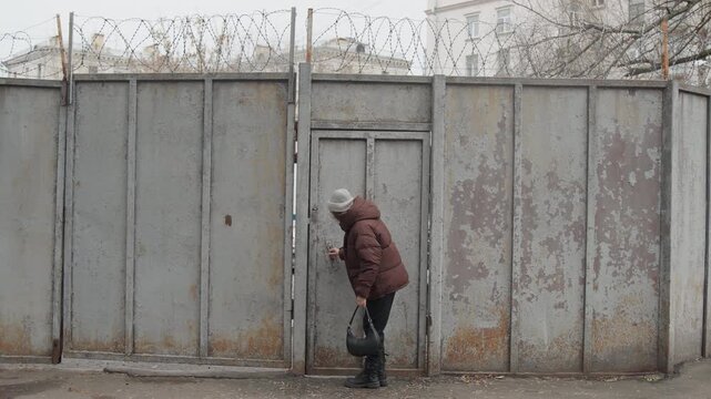 Elderly white woman at rusty gate approaching small metal door, knocking and inspecting lock, opening briefly then walking away, winter coat and knitted hat, carrying small bag, peeling paint