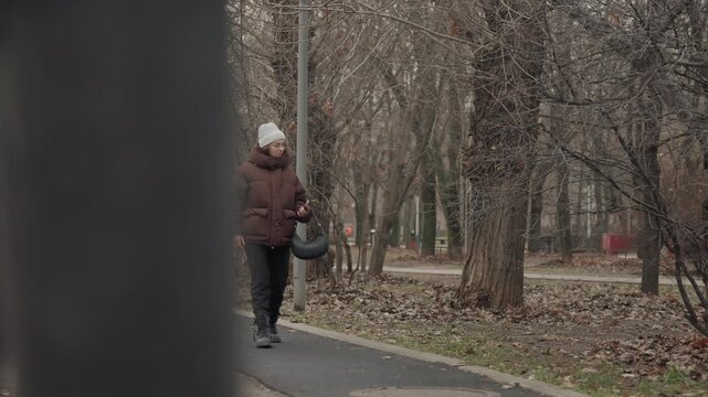 White woman walking through park pathway bundled in puffer coat leafless trees overcast sky carrying leather bag glancing at phone slow cinematic pace introspective mood muted color palette city