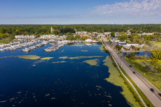 Aerial view of a marina with numerous boats docked in calm water, adjacent to a coastal road and lush green trees in Mears, Michigan, United States.