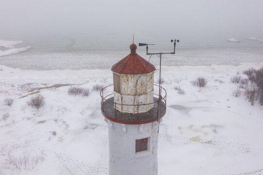 Aerial view of Sturgeon Point Lighthouse with its weathered white tower and rusted red roof, set against a snow-covered landscape and frozen Lake Huron in Harrisville, Michigan, United States.