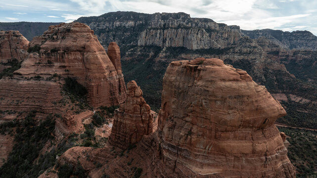Aerial view of the iconic red rock formations featuring towering sandstone cliffs, rugged desert terrain, and sparse vegetation under a cloudy sky in Sedona, Arizona, United States.