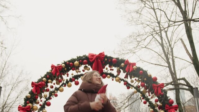 White woman under decorated arch, holding red gnome and looking up at garland and ribbons, bundled in warm coat, tourist vibe on winter boulevard, wide framing with ornamental lights and bare trees