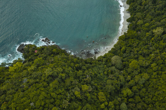Aerial view of Pantai Wedi Ireng featuring dense tropical forest meeting turquoise ocean waters with crashing waves on a rocky coastline in Pesanggaran, East Java, Indonesia.