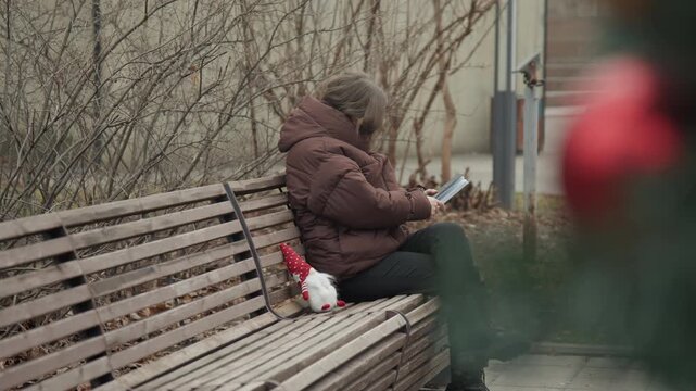 Caucasian woman on bench sketching gnome, blurred red blossom foreground, brown coat and focused profile, intimate courtyard setting with soft bokeh and calm mood, creative pause captured candidly