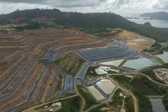 Aerial view of the large open-pit gold and copper mine with terraced slopes, sediment ponds, and surrounding lush hills near the coast in Java, Indonesia.