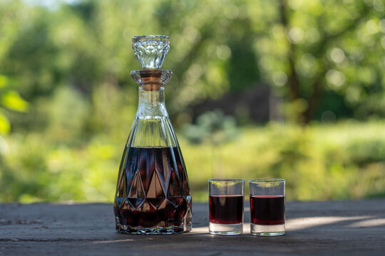 Homemade cherry liqueur in crystal decanter and shot glasses, closeup. Rich dark red homemade cherry liqueur served in vintage crystal decanter and two shot glasses on table in sunny summer garden