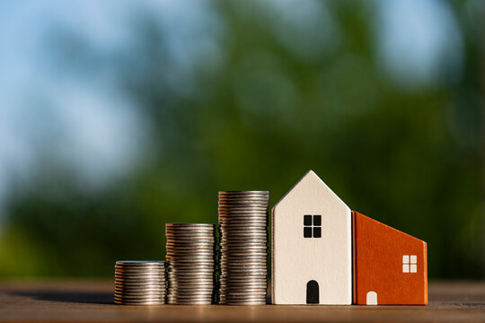 Wooden toy houses next to stacks of coins outdoors. Closeup view of small wooden toy houses standing in line next to growing stacks of silver coins on wooden surface against green nature background