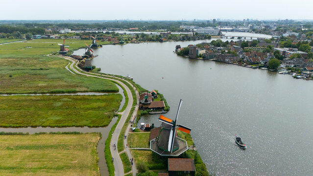 Aerial view of the historic Zaanse Schans windmills along the river with green fields, traditional wooden houses, and a boat cruising on the water in Zaandam, North Holland, Netherlands.