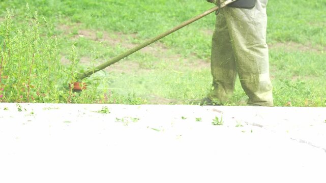 Worker uses a string trimmer on grass. Green lawn fills the background. Dust sprays from the cutting. Concrete path borders the area. Worker wears light trousers