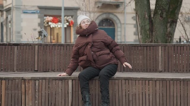 Caucasian woman commuter dazed on bench, regaining consciousness and scanning street for help winter parka and beanie, groggy movements and worried gaze near storefronts and muted traffic, intimate