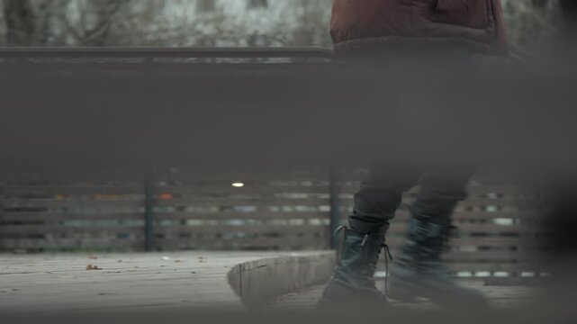 Lowangle boots pacing across curved boardwalk, rhythmic step sequence, overcast park backdrop, railing slats slicing frame, leather creak and scuff, contemplative urban solitude, cinematic tempo
