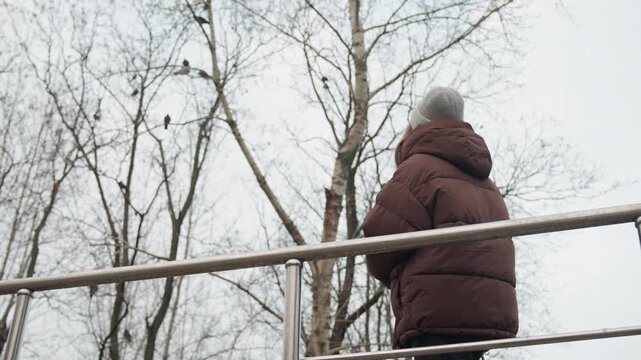 White woman standing on bridge trees, wearing puffer jacket and beanie with back turned toward sky and bare branches, winter overcast mood, contemplative stance and upward gaze suggesting reflection