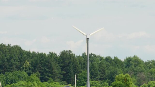 Wind turbine blades turn slowly in the sky. Distant green trees form a natural backdrop. Bird flies overhead near the horizon. Clear sky shows soft cloud patterns