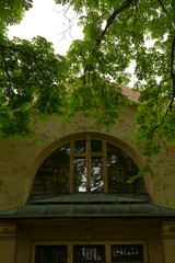 Fototapeta premium Arched window under lush leaves, symmetrical stone arch, glass reflection, textured stucco, quiet leafy foreground and vintage architectural detail.