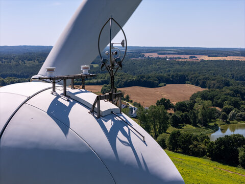 Aerial view of a wind turbine nacelle equipped with meteorological sensors and an anemometer, overlooking a rural landscape with green fields, forests, and a small lake in Poland.