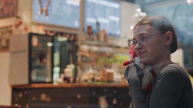 Joyful cafe moment holding small gnome, woman examining plush ornament with delighted grin, shop lights and holiday decor creating warm festive backdrop, candid lifestyle portrait