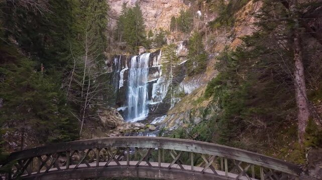 Vue a&eacute;rienne en drone de la grande cascade du cirque de Saint M&ecirc;me, au c&oelig;ur du massif de la Chartreuse, entour&eacute;e d&rsquo;une for&ecirc;t de montagne. Un pont en bois traverse le cours d&rsquo;eau en contrebas de la cas