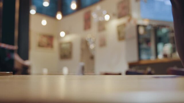 Caucasian hand picking pastry from closed cafe table, small ribbonwrapped sweet on wooden surface, delicate grasp and reaching fingers, empty dining room with cordon and warm lights, intimate closeup
