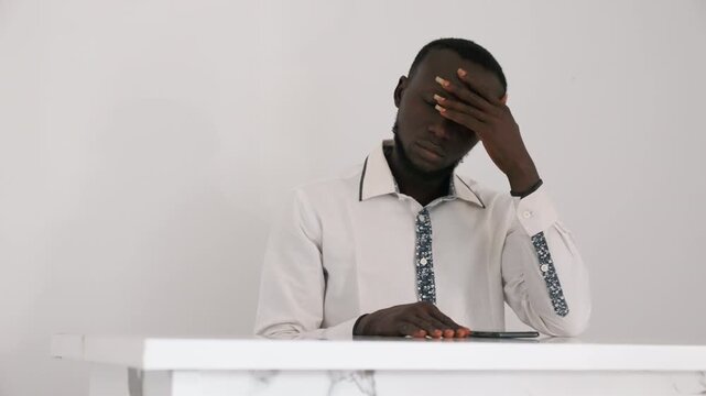 Black man sitting at table with hand on forehead, white shirt and patterned tie, minimal interior, phone on table, tense posture suggesting worry about date and private disappointment in quiet scene.
