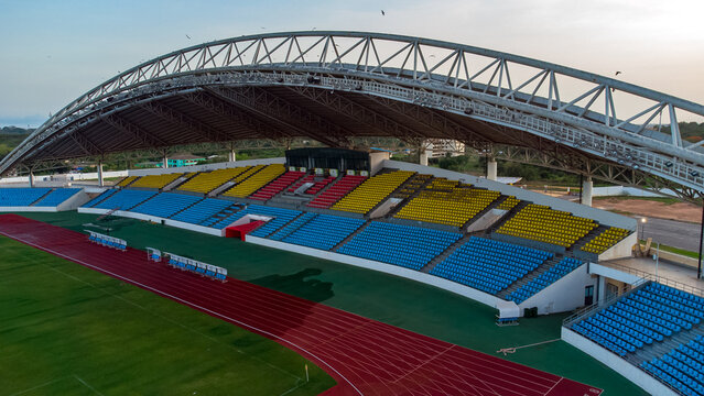 Aerial view of Cape Coast Sports Stadium with its large arched roof, vibrant blue, yellow, and red seating, and a running track in Cape Coast, Central Region, Ghana.