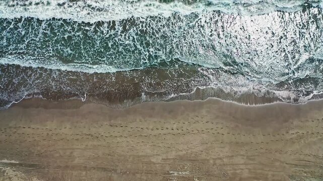 Sea waves crashing on beach vertical aerial view in a top down angle. Dramatic drone shot capturing turquoise ocean foam washing onto sandy shoreline.