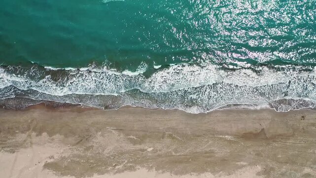 Ocean waves hitting sandy beach aerial vertical view with dynamic movement. Stunning drone camera capturing vibrant blue water washing over golden shore.