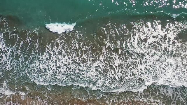 Sea waves crashing on beach vertical aerial view in a top down angle. Dramatic drone shot capturing turquoise ocean foam washing onto sandy shoreline.