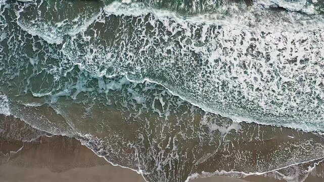 Ocean waves hitting sandy beach aerial vertical view with dynamic movement. Stunning drone camera capturing vibrant blue water washing over golden shore.