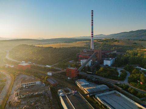 Aerial view of the Kronospan woodworks facility with its tall chimney, factory buildings, and conveyor belt set against rolling hills in Zvolen, Banskobystricky kraj, Slovakia.