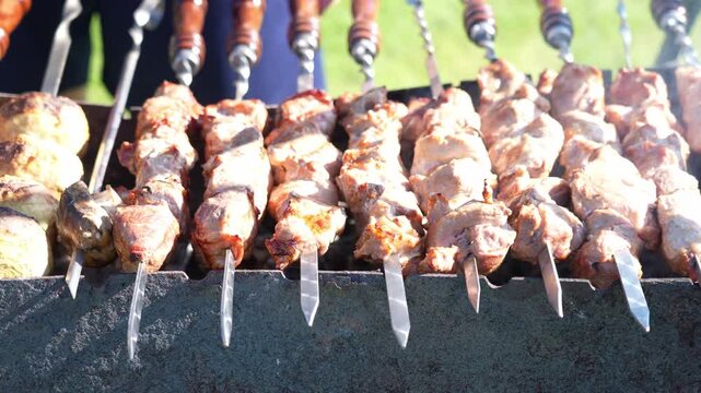 Close-up of skewered meat on a grill. Meat is partially cooked, golden brown in spots. Some skewers show charred edges. Person adjusts the skewers. Bright sunny day outdoors. Ideal for food, cooking