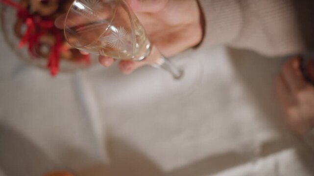 Hand holding crystal flute over table, soft warm light, bowl of ribboned pretzels and scattered oranges, beige sweater sleeve, empty glass catching highlights, slow cinematic gesture conveying cozy